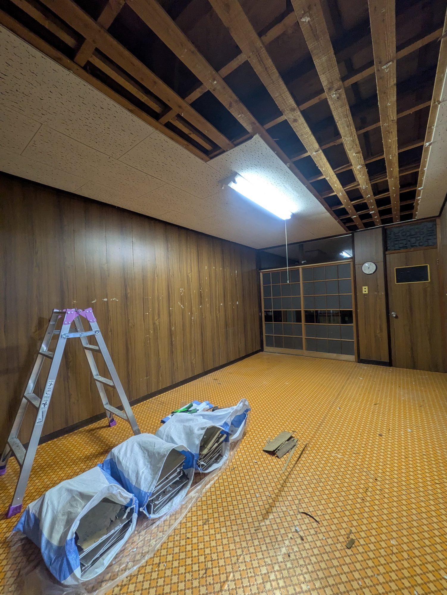 天井板を剥がし、梁がむき出しになった和室の様子。奥に建具、手前に床と養生された道具一式が見える